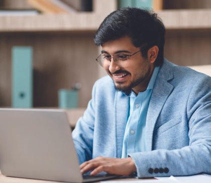 indian-man-wearing-light-blue-jacket-sits-desk-modern-office-smiling-as-he-works-his-001