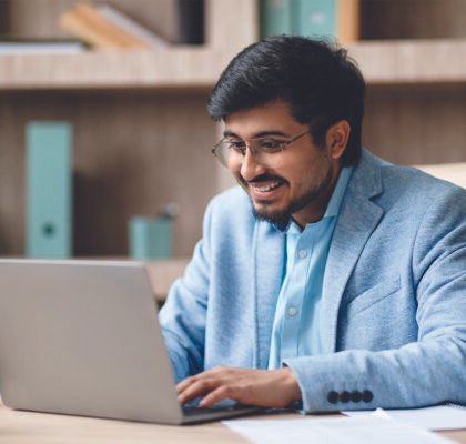 indian-man-wearing-light-blue-jacket-sits-desk-modern-office-smiling-as-he-works-his-001